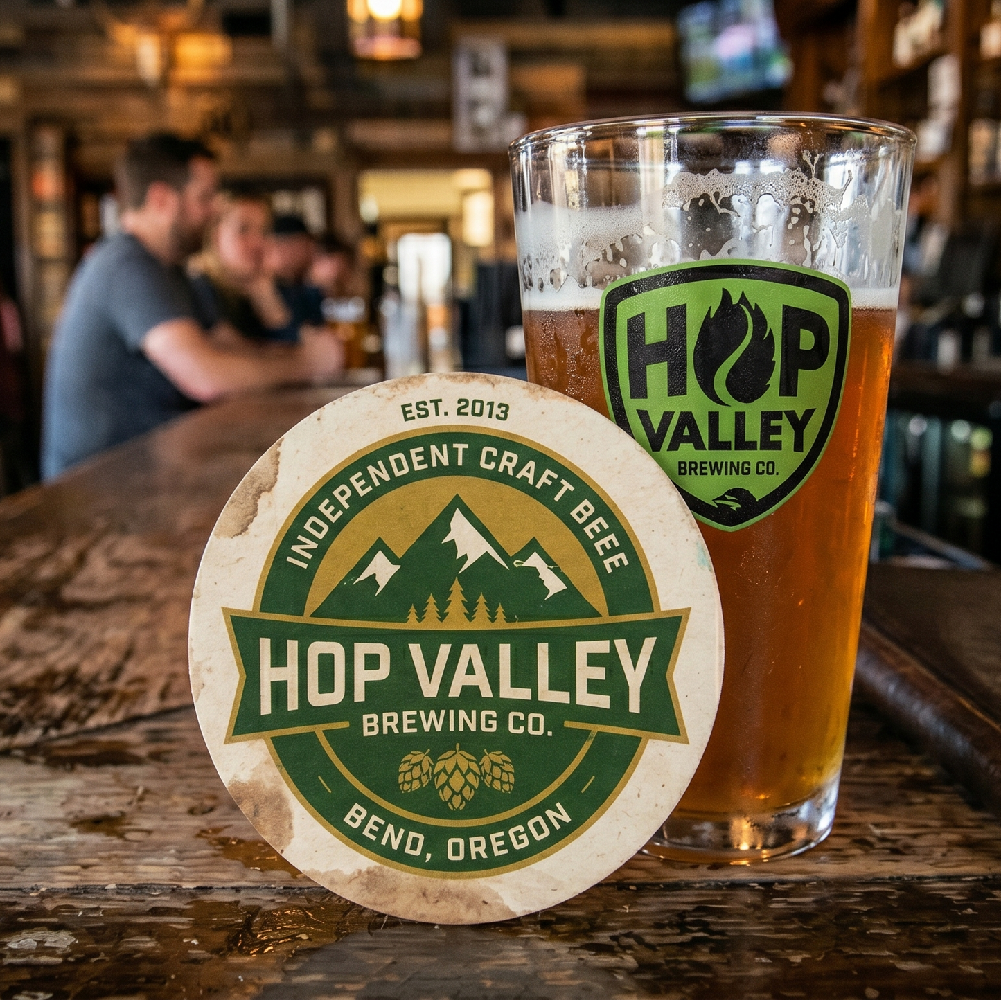 Glass of beer with a coaster from Hop Valley Brewing Co. on a bar counter with patrons in the background.